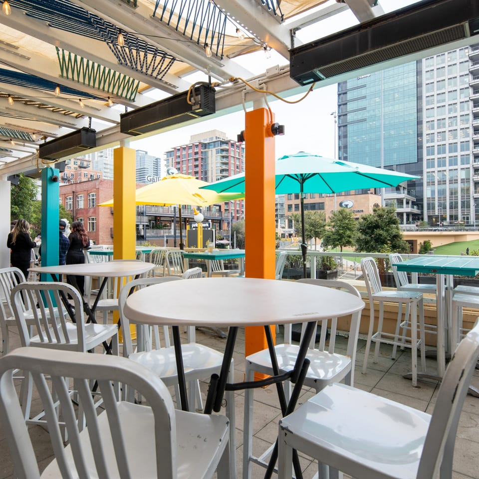 Fairweather bar's outdoor patio with white tables and chairs, colorful umbrellas, and city buildings in the background.