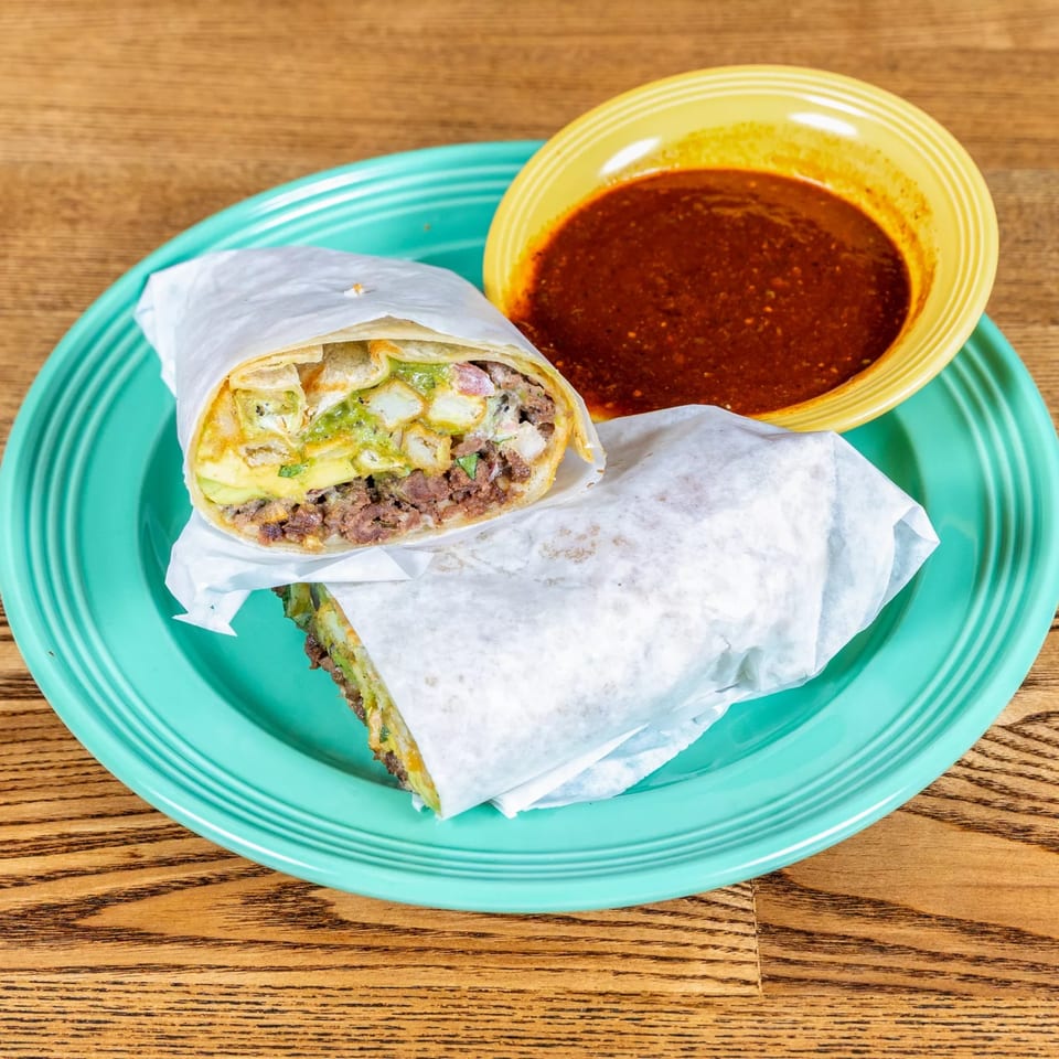 Two halves of a burrito with meat, cheese, and guacamole, served with a side of red sauce.