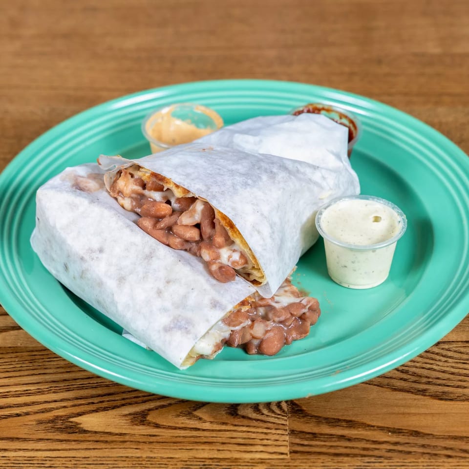 A Fairweather bean and cheese burrito, wrapped in paper, on a teal plate with two dipping sauces.